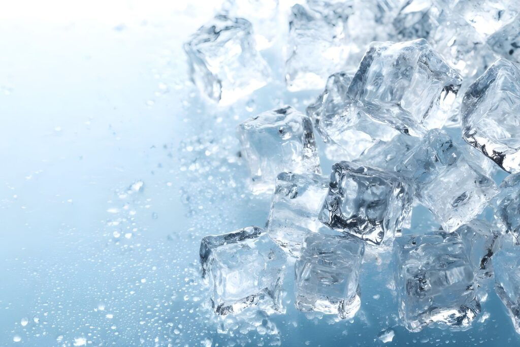 Close-up view of transparent ice cubes piled and scattered across a cold light blue surface with sparkling frost and water droplets, creating a fresh and chilled atmosphere.
